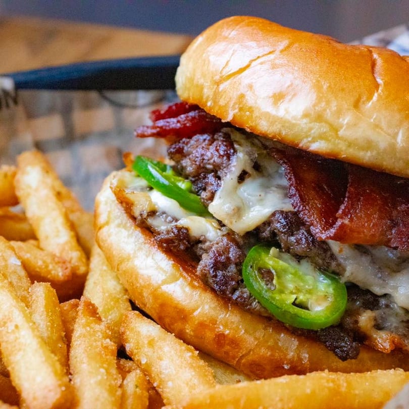 Close-up of a BBQ King Smokehouse burger with bacon, melted cheese, and jalapeños, served with fries.